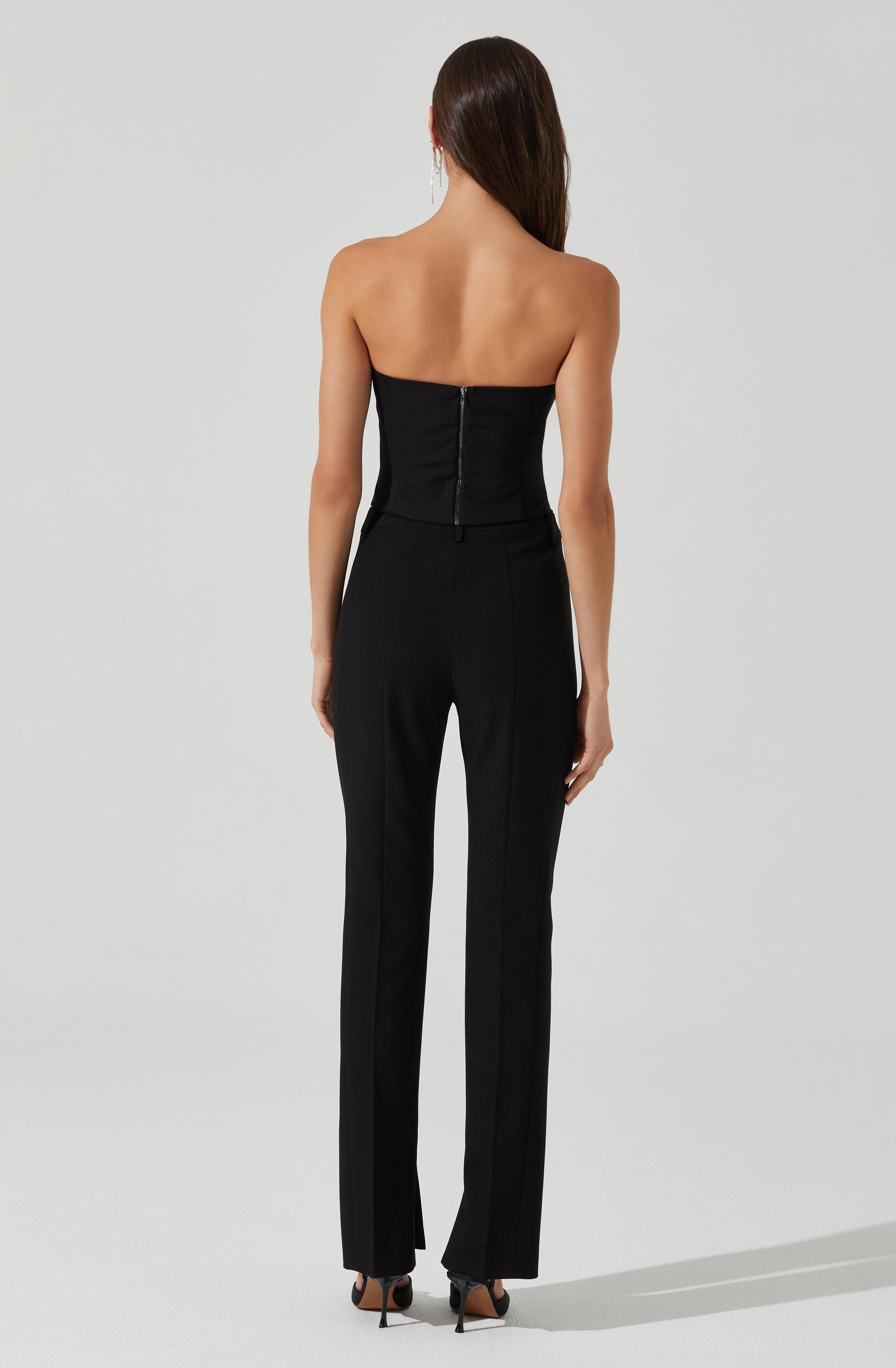 A woman is wearing a strapless black top with a zipper at the back, paired with black, straight-leg trousers. She is also wearing black high-heeled shoes.