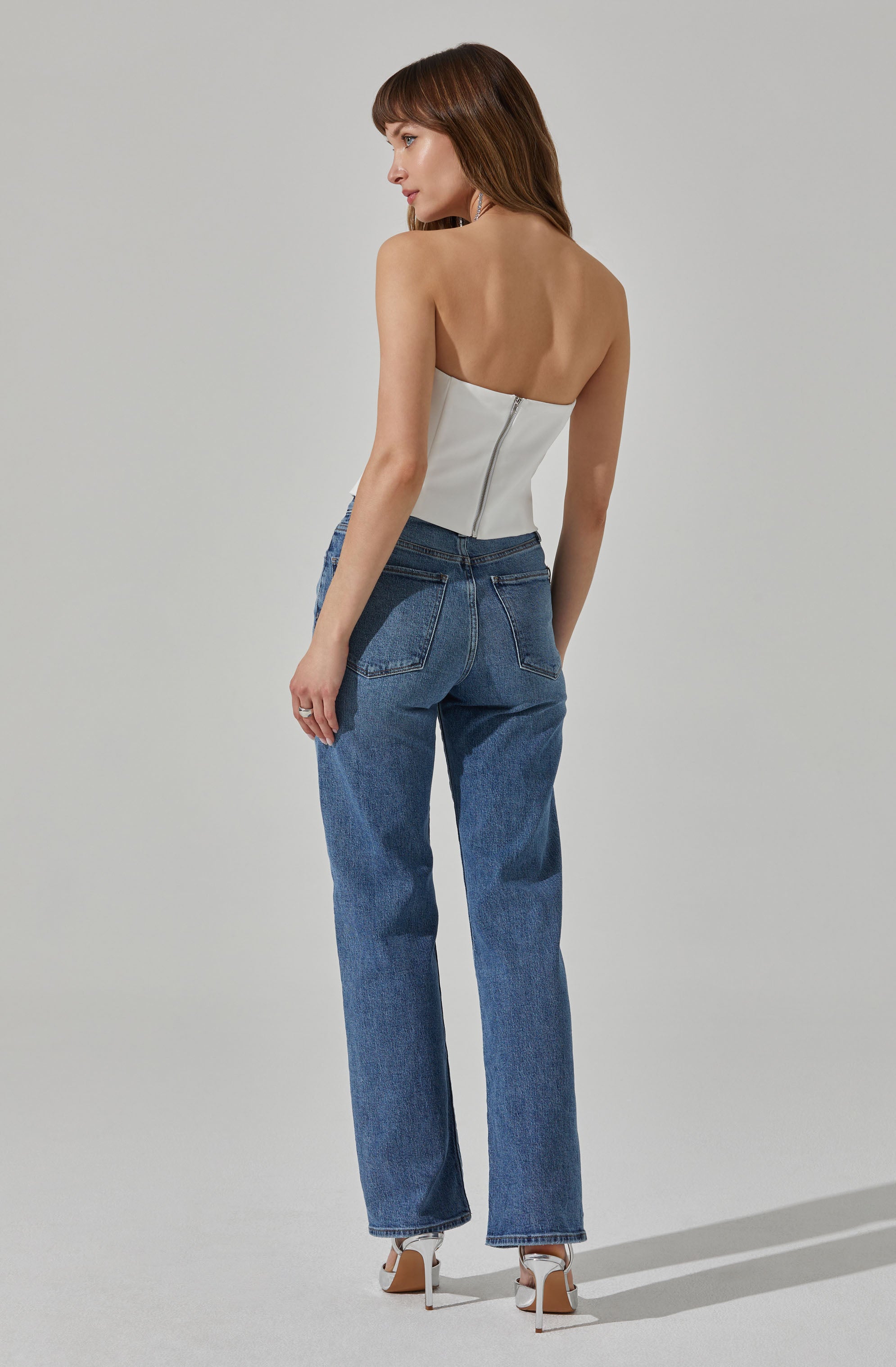 A woman is wearing a white, strapless top with a back zipper opening, paired with high-waisted blue jeans. She is also wearing white high-heeled shoes.