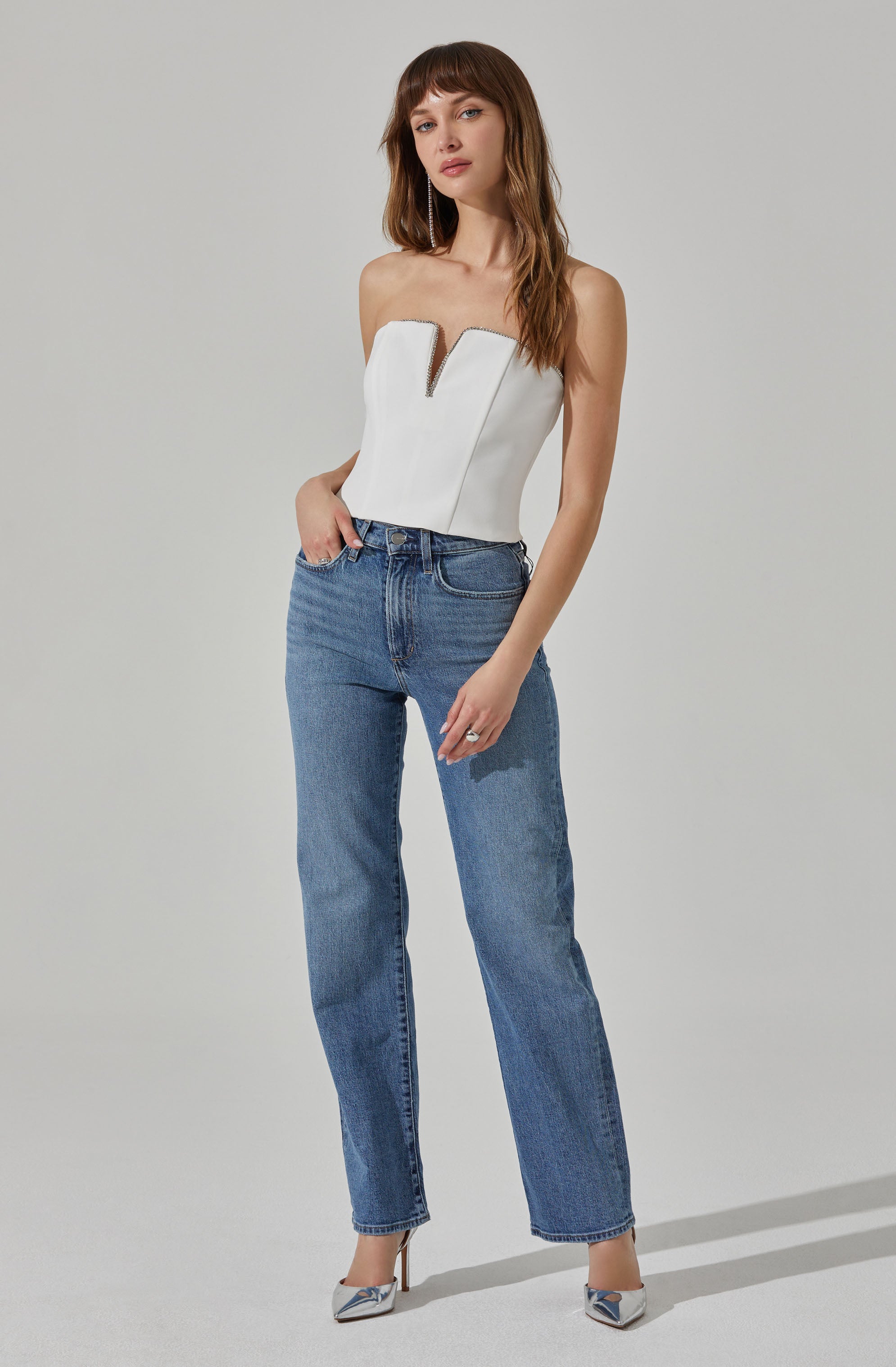 A woman is wearing a strapless white top with a front zipper detail, paired with high-waisted blue jeans that have a relaxed, straight-leg fit. She completes her outfit with pointed silver shoes.