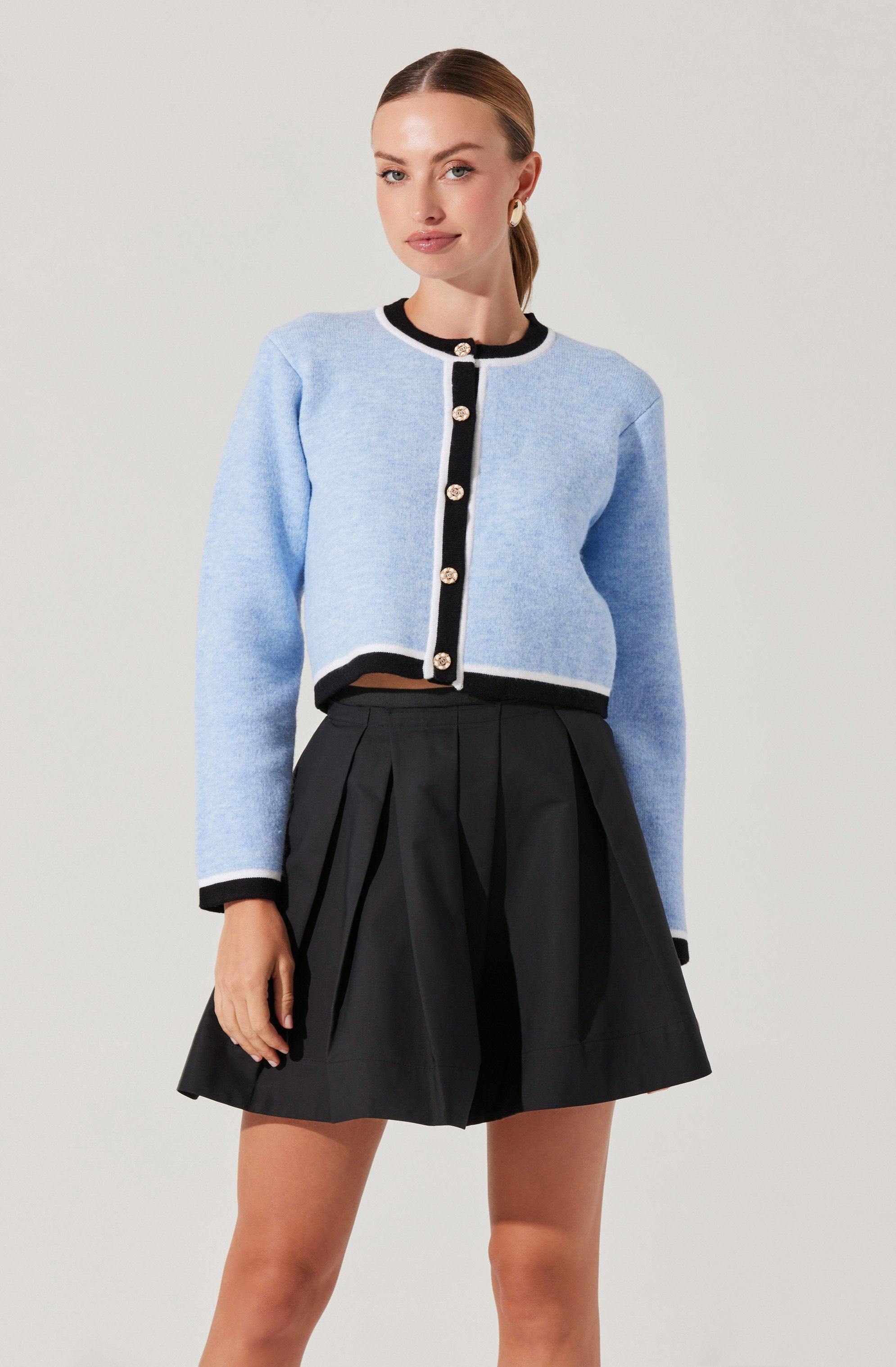 A woman is wearing a light blue cropped cardigan with black trim and large decorative buttons down the front. The cardigan has long sleeves, and the black trim is present along the collar, cuffs, and hem. She is also wearing a high-waisted black pleated skirt that reaches above her knees.