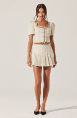 A woman is wearing a coordinated outfit made of a light-colored, textured fabric with a checkered pattern. The top is a cropped jacket with short puff sleeves, a rounded neckline, and decorative trim along the edges and the front opening. It has button closures down the front. The matching skirt is high-waisted, pleated, and short, ending well above the knees. She is also wearing black pointed-toe heels.