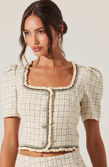 A woman is wearing a short-sleeved, cream-colored, textured tweed jacket with black trim along the neckline, front opening, and hem. The jacket features a rounded square neckline, puffed shoulders, and decorative pearl buttons.