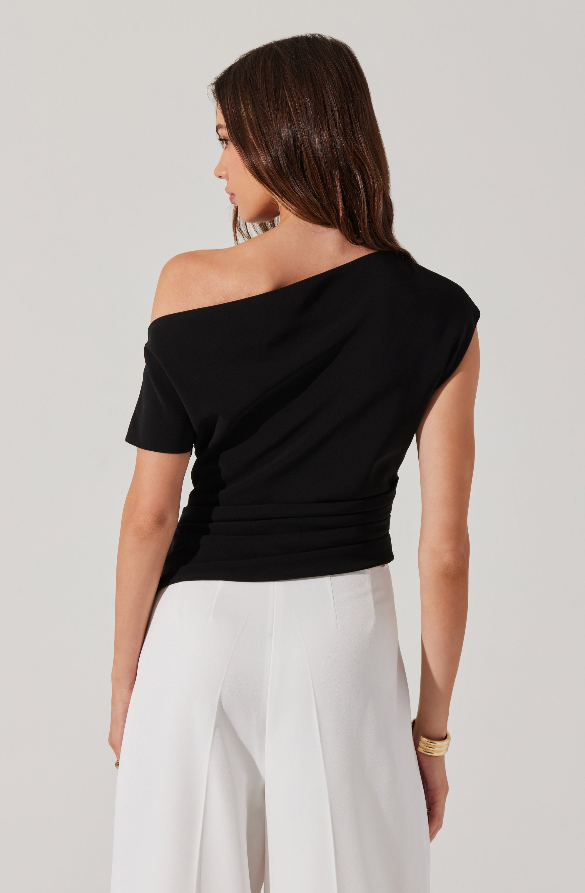 A woman is wearing a black, off-the-shoulder, short-sleeved top with a draped design and a fitted waist. She pairs it with high-waisted white wide-leg pants and accessorizes with a gold bracelet on her right wrist.
