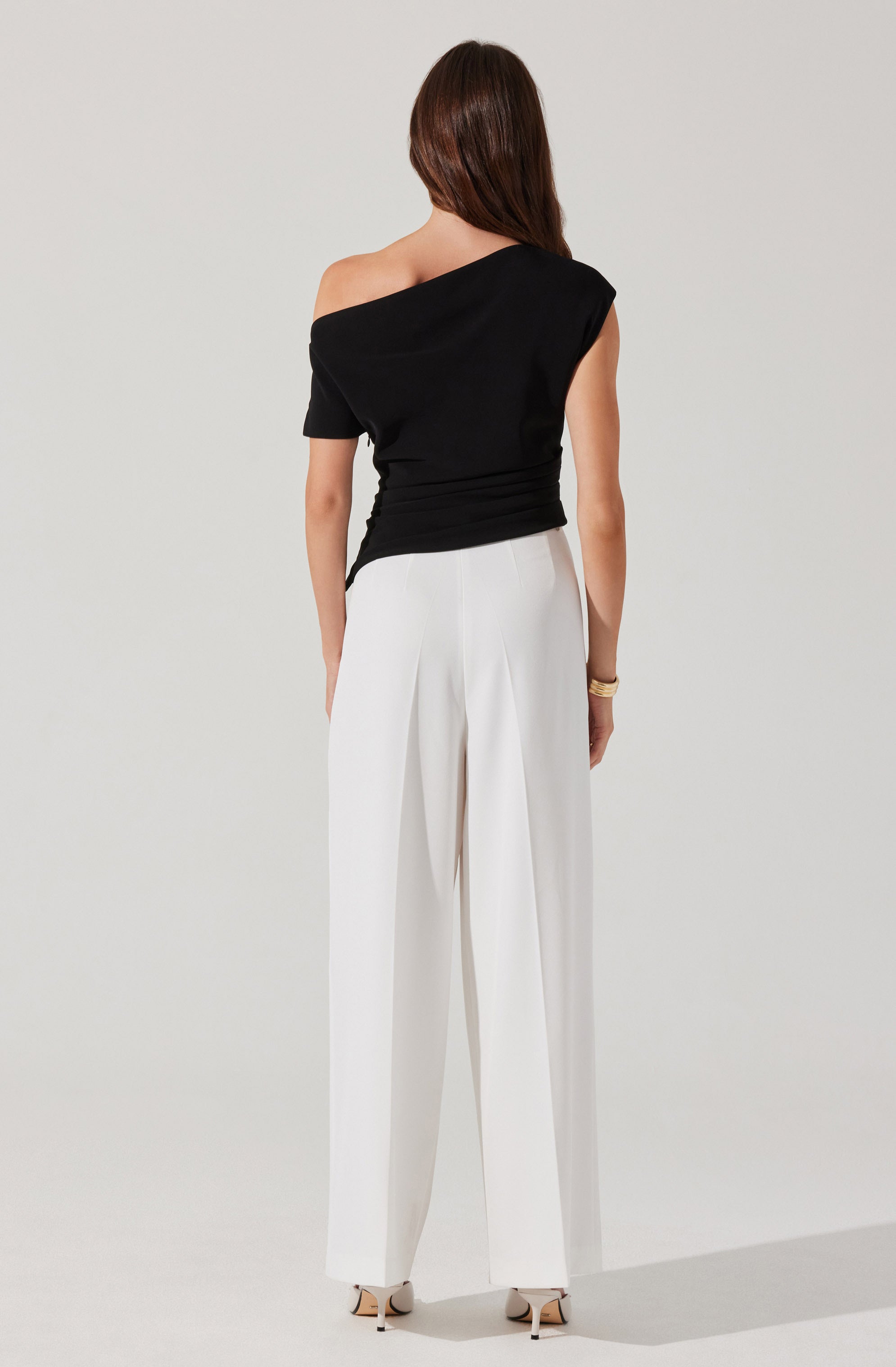 A woman is wearing a black, off-the-shoulder top with short sleeves and a relaxed fit. She pairs it with high-waisted, wide-leg white trousers that have pleats at the front. The outfit is complemented by a bracelet on her right wrist and beige high-heeled shoes.