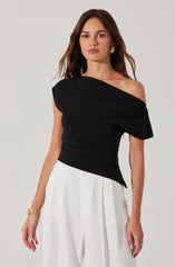 The woman is wearing a black, asymmetrical top with one shoulder exposed and the other covered by a sleeve. The top is fitted with gathered or ruched details around the waist. She is also wearing white, wide-legged trousers. She has gold earrings, a gold bracelet, and a gold ring as accessories.