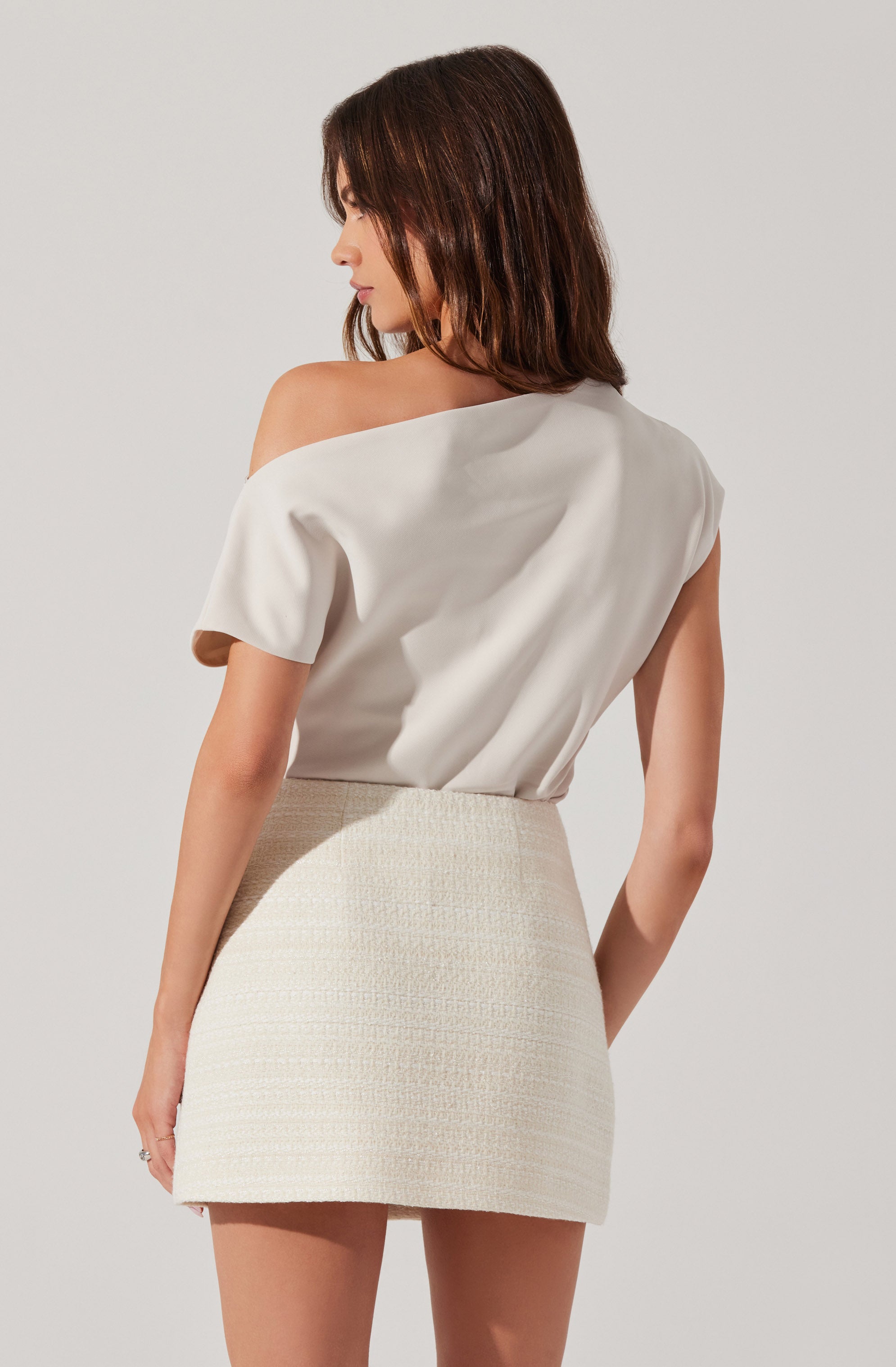 A woman is wearing an off-the-shoulder, short-sleeved white top made of a smooth fabric, paired with a high-waisted, textured cream-colored mini skirt.