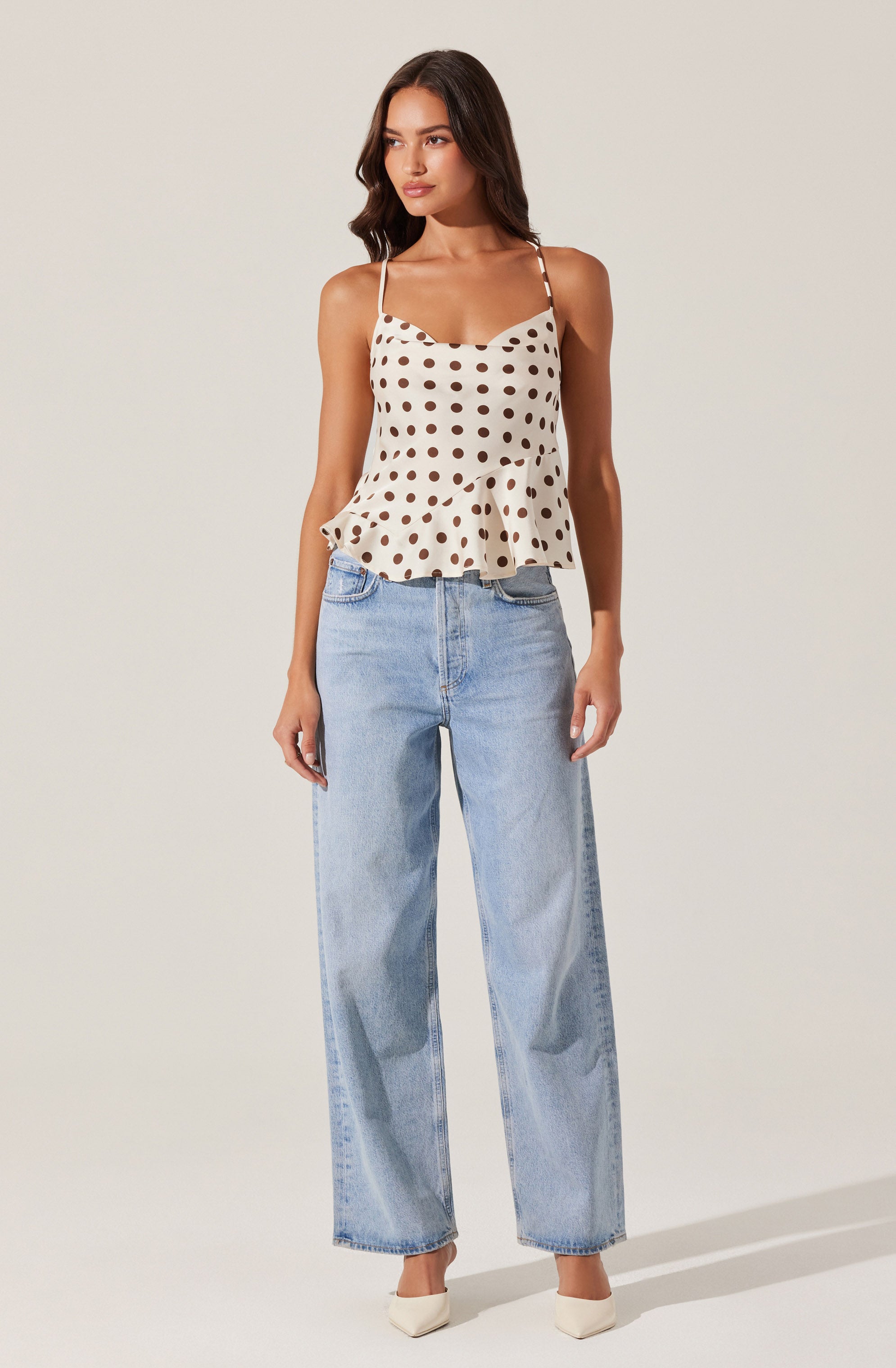 A woman is wearing a cream-colored sleeveless top with dark brown polka dots, featuring thin straps and a ruffled hem. She has paired it with light blue, high-waisted, wide-leg jeans. She is also wearing white pointed-toe shoes.