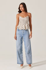 A woman is wearing a cream-colored sleeveless top with dark brown polka dots, featuring thin straps and a ruffled hem. She has paired it with light blue, high-waisted, wide-leg jeans. She is also wearing white pointed-toe shoes.