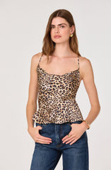 A woman is wearing a sleeveless, leopard-print top with spaghetti straps and a ruffled hem, paired with high-waisted dark blue jeans. She has gold hoop earrings and a gold bracelet.
