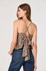 A woman is wearing a sleeveless top with thin, leopard-print spaghetti straps and a large bow tied at the back. The top features a leopard print pattern with shades of tan, brown, and black. She pairs it with high-waisted dark blue jeans.