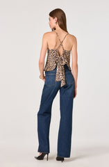 The woman is wearing a sleeveless, leopard print top with thin spaghetti straps that crisscross at the back and tie into a bow. She is also dressed in high-waisted blue jeans with large back pockets, paired with black high-heeled shoes.