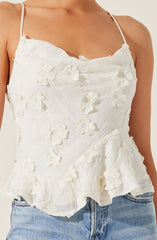 A woman is wearing a sleeveless, white, floral textured top with thin spaghetti straps. The top has a slightly ruffled hemline and features 3D floral appliqué details throughout. She pairs it with light blue denim jeans.