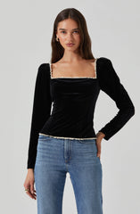 A young woman is wearing a black velvet long-sleeve top with a square neckline that is decorated with a white ruffled trim. The top fits snugly and has slight puffiness at the shoulders. She pairs it with light blue high-waisted jeans.