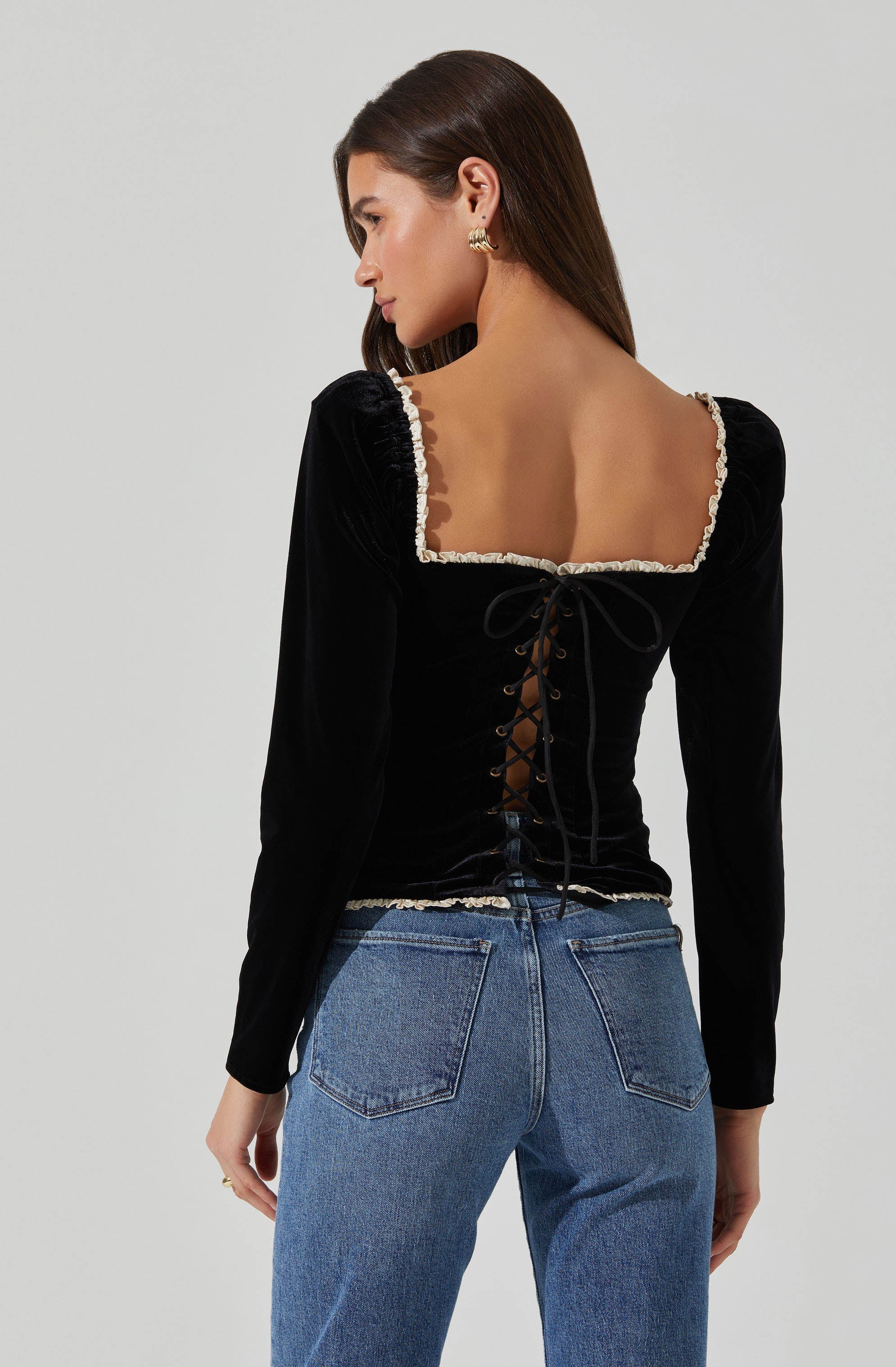 A woman wearing a fitted black velvet top with long sleeves and a square neckline trimmed with lace. The back of the top features a lace-up detail with black ties. She is also wearing high-waisted blue jeans with visible stitching and back pockets.