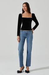 A woman is wearing a black, long-sleeved, velvet top with a square neckline accented by a decorative trim. She has paired the top with light blue, high-waisted, straight-leg jeans and black high-heeled shoes with bows on the front.