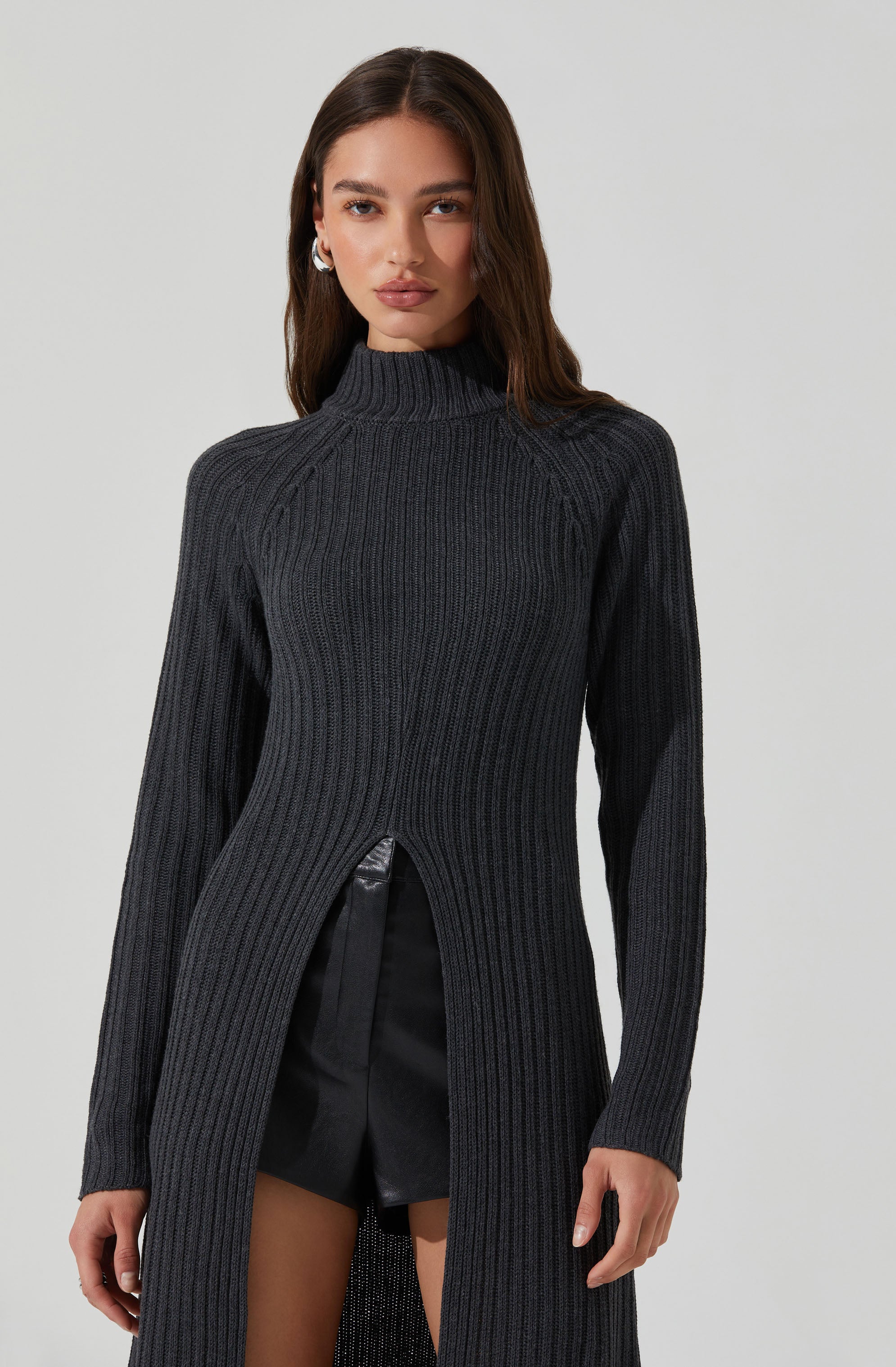 A woman is wearing a long, dark gray, ribbed knit sweater with a high neckline. The sweater features a front slit from the waist down, revealing black shorts underneath. The sweater has a fitted silhouette with long sleeves.