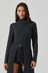 A woman is wearing a long, dark gray, ribbed knit sweater with a high neckline. The sweater features a front slit from the waist down, revealing black shorts underneath. The sweater has a fitted silhouette with long sleeves.