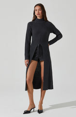 The model is wearing a long, dark gray, ribbed knit sweater with a high neckline. The sweater has a body-hugging fit on the upper part and features a front slit that reveals black shorts underneath. She is also wearing black pointed-toe high heels with decorative embellishments.