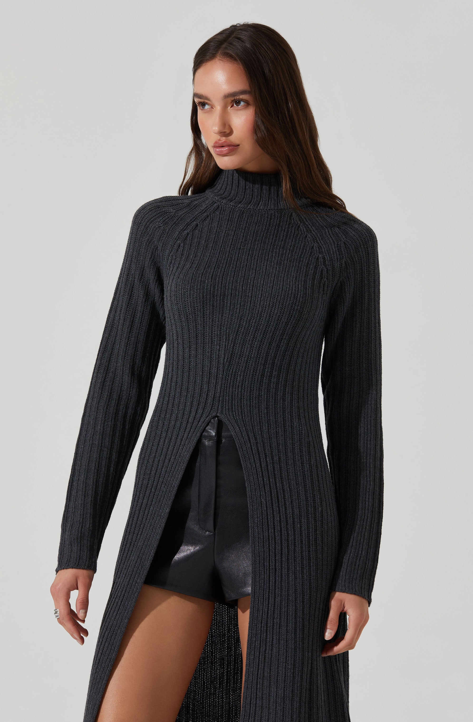 A woman is wearing a long, dark gray, ribbed knit sweater with a high neckline. The sweater has a slit from the waist down, revealing black leather shorts underneath. The sweater's design includes vertical ribbing and a semi-open front from the waist down.