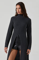 A woman is wearing a long, dark gray, ribbed knit sweater with a high neckline. The sweater has a slit from the waist down, revealing black leather shorts underneath. The sweater's design includes vertical ribbing and a semi-open front from the waist down.