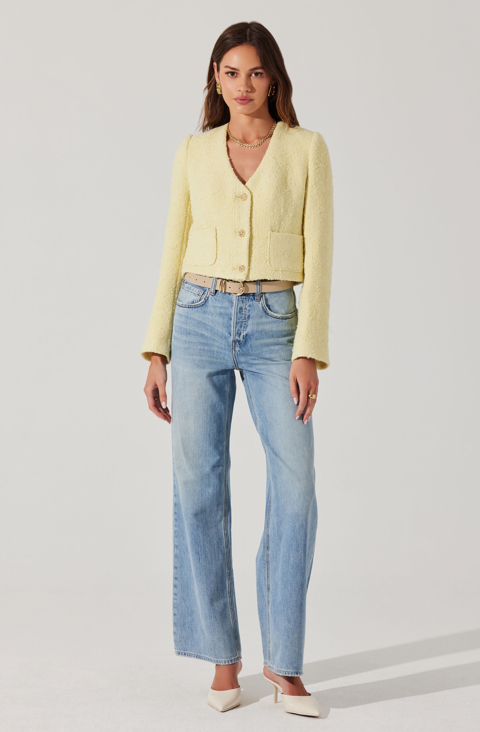 A woman is wearing a light yellow textured, cropped cardigan with three large buttons down the front and two patch pockets at the waist. She has paired it with light blue, high-waisted, straight-leg jeans and white pointed-toe heels. She accessorizes with gold hoop earrings, a gold chain necklace, and a beige belt with a gold buckle.