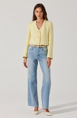 A woman is wearing a light yellow textured, cropped cardigan with three large buttons down the front and two patch pockets at the waist. She has paired it with light blue, high-waisted, straight-leg jeans and white pointed-toe heels. She accessorizes with gold hoop earrings, a gold chain necklace, and a beige belt with a gold buckle.