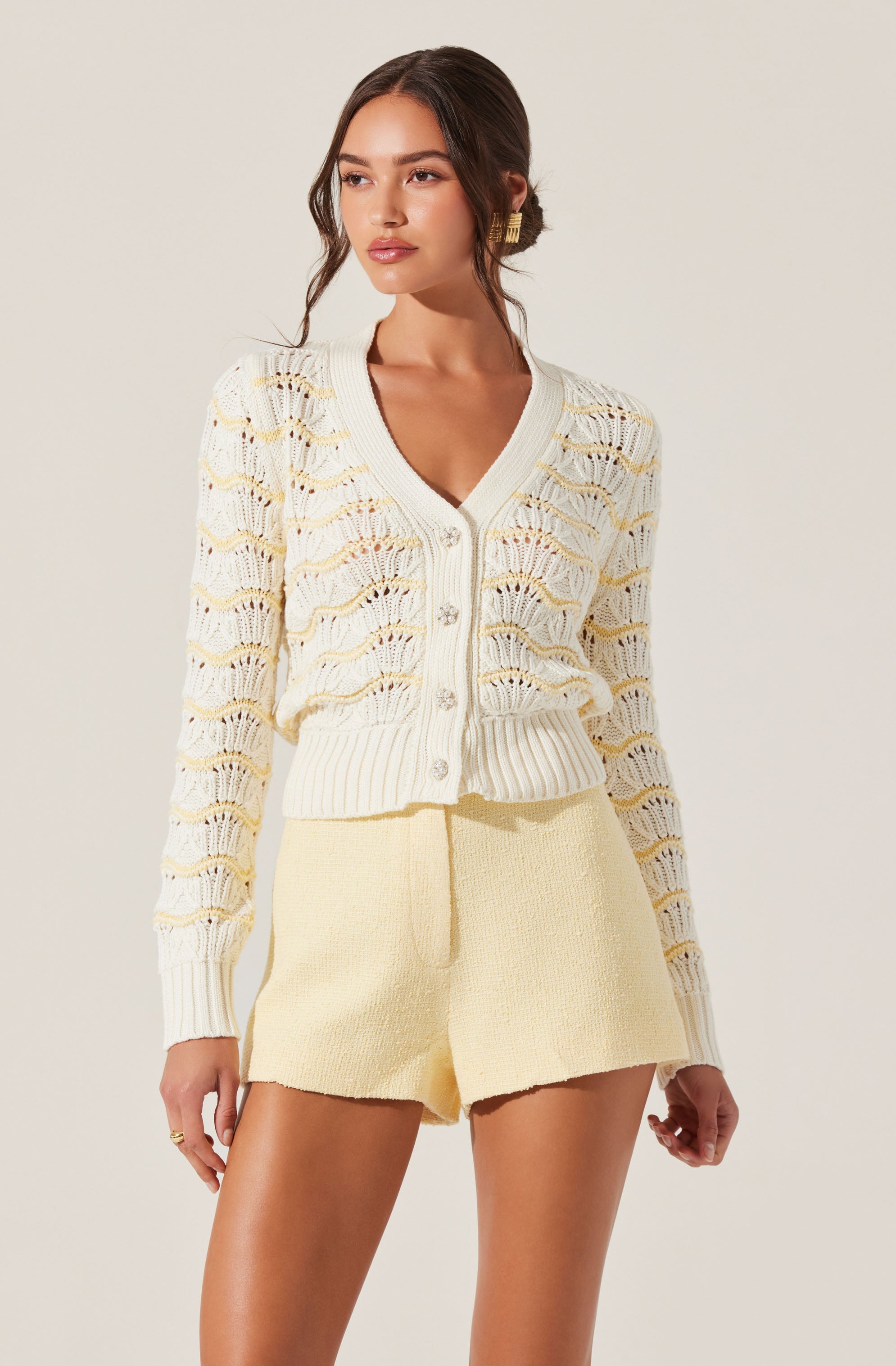 A woman is wearing a cream-colored, open-knit cardigan with a button-up front, featuring a scalloped design and ribbed hem and cuffs. She pairs it with high-waisted, cream-colored textured shorts.