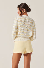 A woman is wearing a cream-colored, open-knit sweater with a scalloped wave pattern and long sleeves, paired with matching high-waisted shorts. She has her dark hair styled in an elegant low bun and is accessorized with large, gold, geometric earrings.