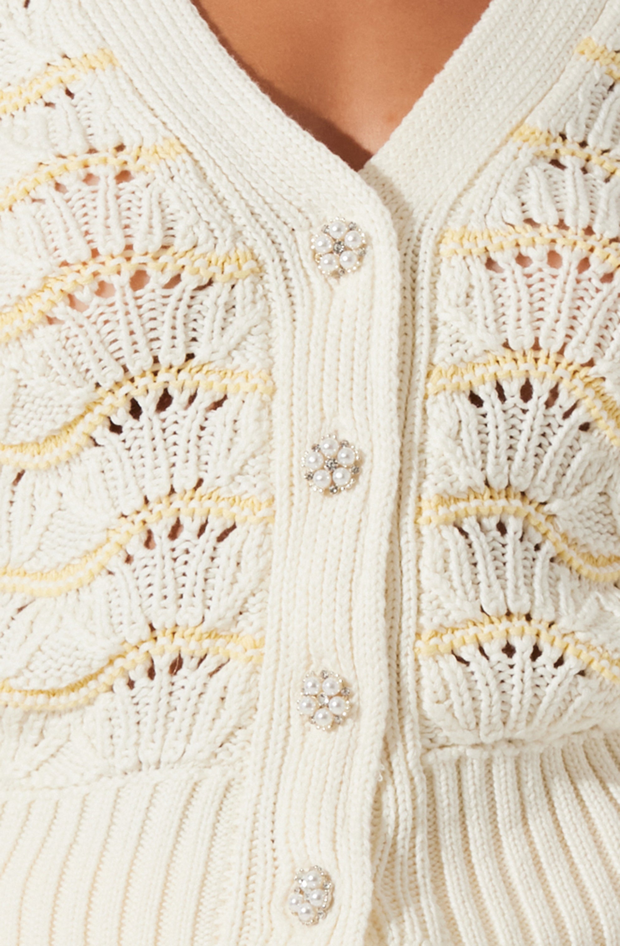An off-white, knitted cardigan featuring decorative pearl and rhinestone buttons. The cardigan has a textured, open-weave pattern with horizontal stripes of different knit styles. The buttons are round with a floral design made of small pearls and rhinestones. The neckline is a V-shape.