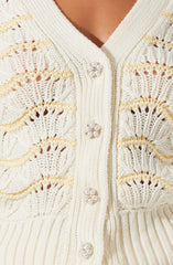 An off-white, knitted cardigan featuring decorative pearl and rhinestone buttons. The cardigan has a textured, open-weave pattern with horizontal stripes of different knit styles. The buttons are round with a floral design made of small pearls and rhinestones. The neckline is a V-shape.