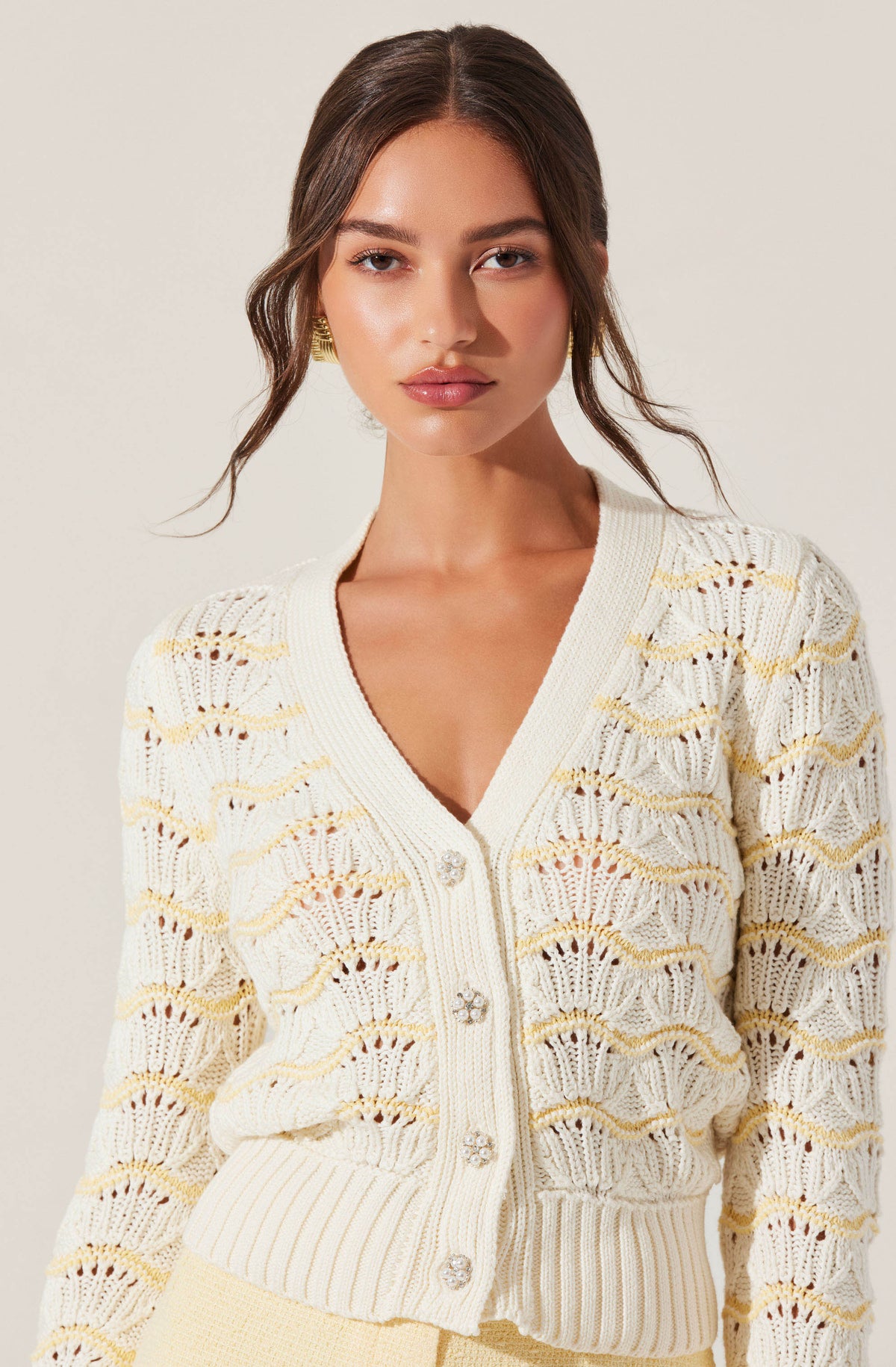 A woman is wearing a cream-colored, open-knit cardigan with a scalloped pattern and ribbed cuffs and hem. The cardigan features decorative rhinestone buttons down the front.