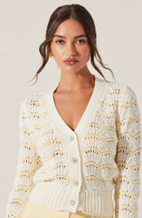 A woman is wearing a cream-colored, open-knit cardigan with a scalloped pattern and ribbed cuffs and hem. The cardigan features decorative rhinestone buttons down the front.