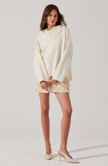 A woman is wearing an oversized, cream-colored sweater with long sleeves and a wide, folded neckline. She pairs it with a short, textured cream-colored skirt that has an intricate, fringed design. Her footwear consists of light-colored pointed-toe heels with ankle straps.