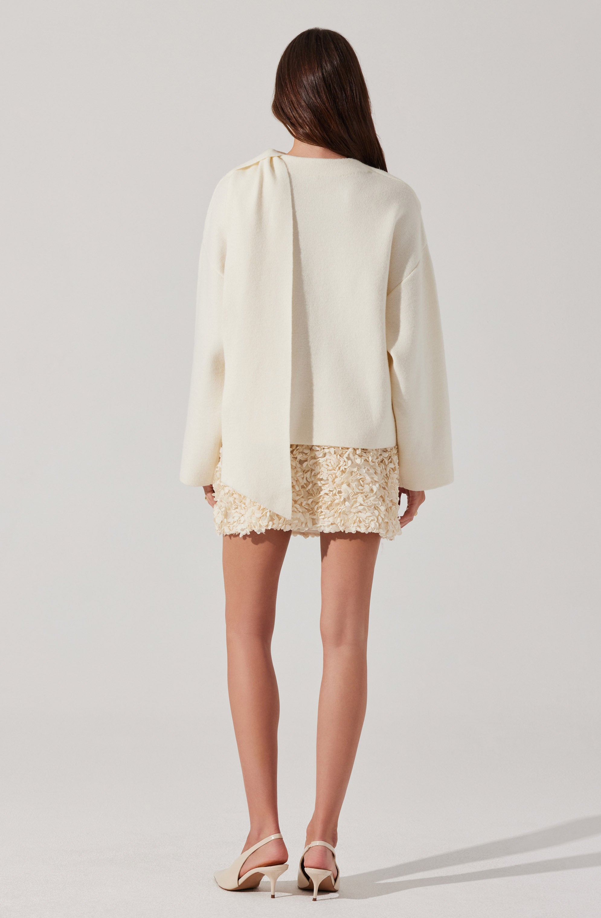 A woman is wearing a loose-fitting, long-sleeved white top with a gathered detail at the shoulder. She has paired it with a short, textured cream-colored skirt that features a floral or ruffled appliqué pattern. She is also wearing white open-toe high-heeled sandals.