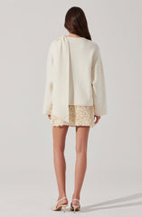 A woman is wearing a loose-fitting, long-sleeved white top with a gathered detail at the shoulder. She has paired it with a short, textured cream-colored skirt that features a floral or ruffled appliqué pattern. She is also wearing white open-toe high-heeled sandals.