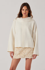 A woman is wearing an oversized, off-white top with a wide, rounded collar and long sleeves. She pairs it with a short, textured cream-colored skirt that has a floral, ruffled pattern.