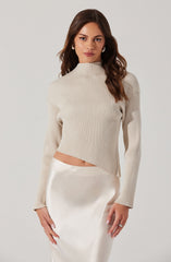 A woman is wearing a cream-colored, ribbed, long-sleeve, high-neck sweater with a cropped fit. She pairs it with an elegant, satin, ivory-colored skirt that has a smooth and shiny texture.