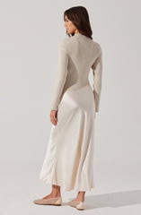 A woman is wearing a light beige, fitted, ribbed long-sleeve sweater with a high neckline. She pairs it with a long, flowing, satin-like cream-colored skirt that reaches her ankles and features a subtle slit on the side. She completes the outfit with beige, pointed-toe ballet flats that have a textured woven pattern.
