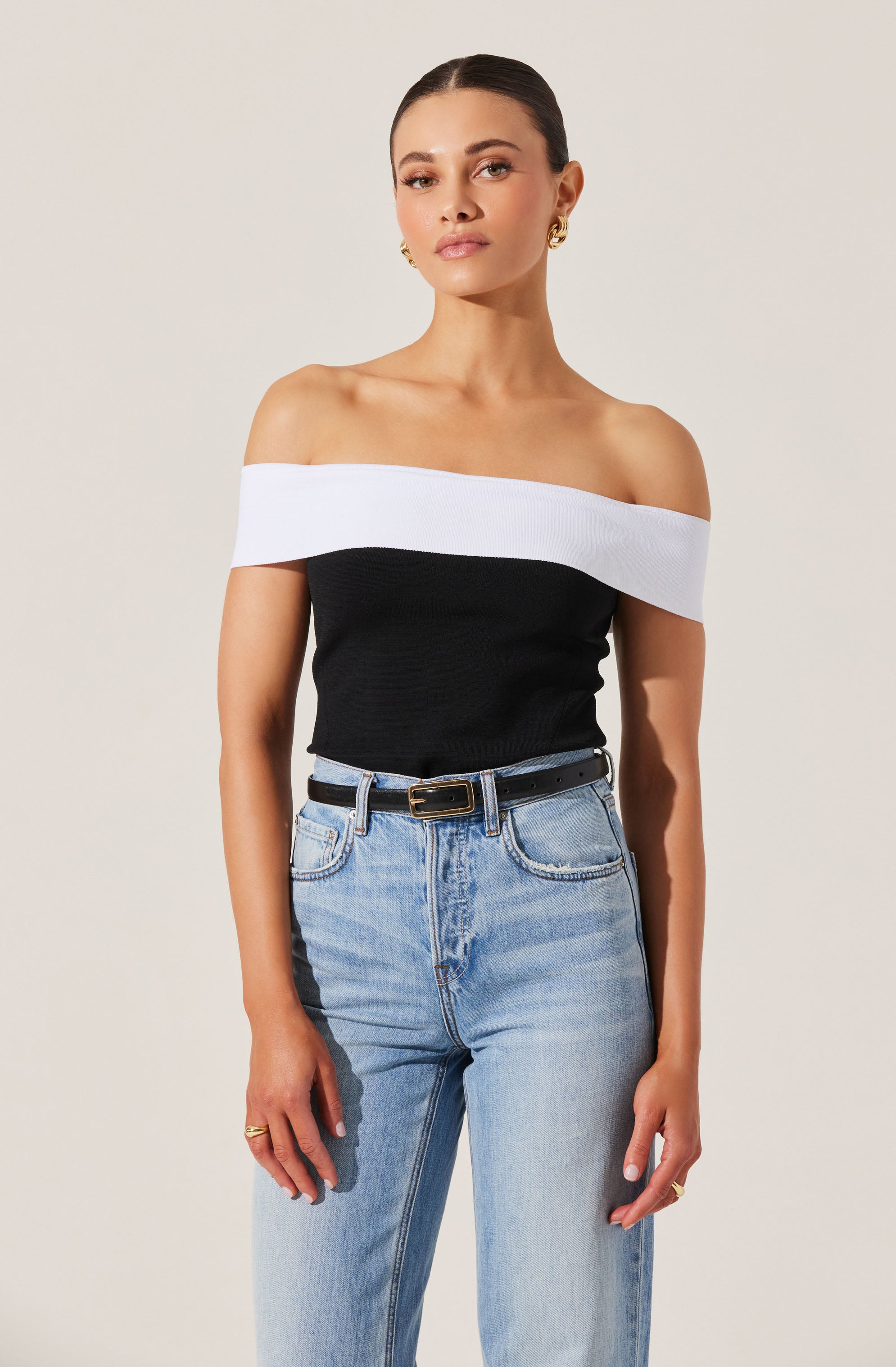 A woman is wearing a black off-the-shoulder top with a wide white band across the neckline, paired with light blue high-waisted jeans and a black belt with a gold buckle. She has gold earrings and rings on her fingers.