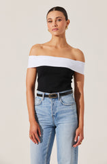 A woman is wearing a black off-the-shoulder top with a wide white band across the neckline, paired with light blue high-waisted jeans and a black belt with a gold buckle. She has gold earrings and rings on her fingers.