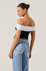 A woman is wearing an off-the-shoulder top with a wide white band around the shoulders, and the main part of the top is black. She has paired it with high-waisted light blue jeans that have a slightly loose fit. She is also wearing gold earrings and rings. Her hair is styled in a sleek bun.
