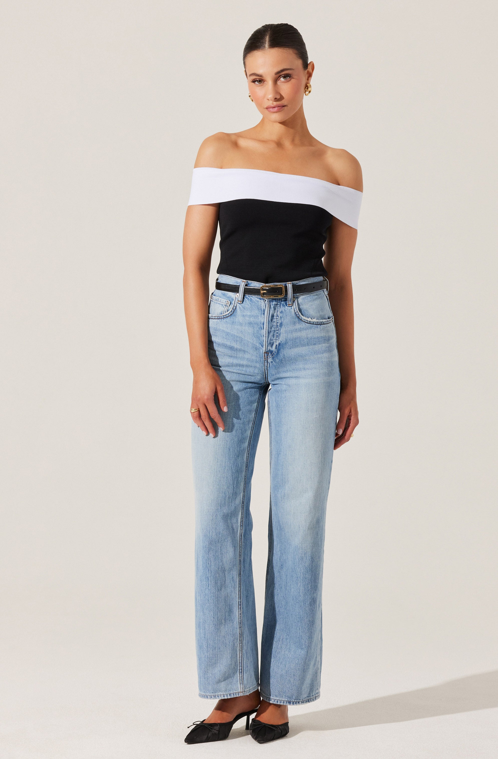 A woman is wearing an off-the-shoulder top with a wide white band across the shoulders and a black body. She has paired it with high-waisted, light-wash denim jeans that have a relaxed, straight-leg fit. The jeans are secured with a black belt with a gold buckle. She is also wearing black pointed-toe heels.