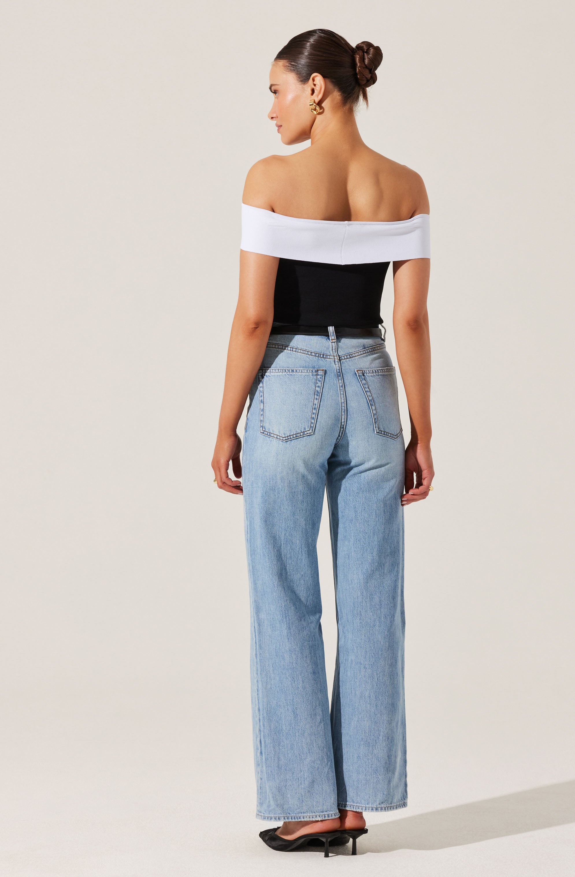 A woman is wearing an off-the-shoulder top with white fabric around the shoulders and black fabric covering the torso. She is also wearing high-waisted, light-wash, wide-leg jeans with visible stitching and back pockets. The outfit is completed with black heeled shoes. Her hair is styled in a neat bun, and she has on gold hoop earrings.
