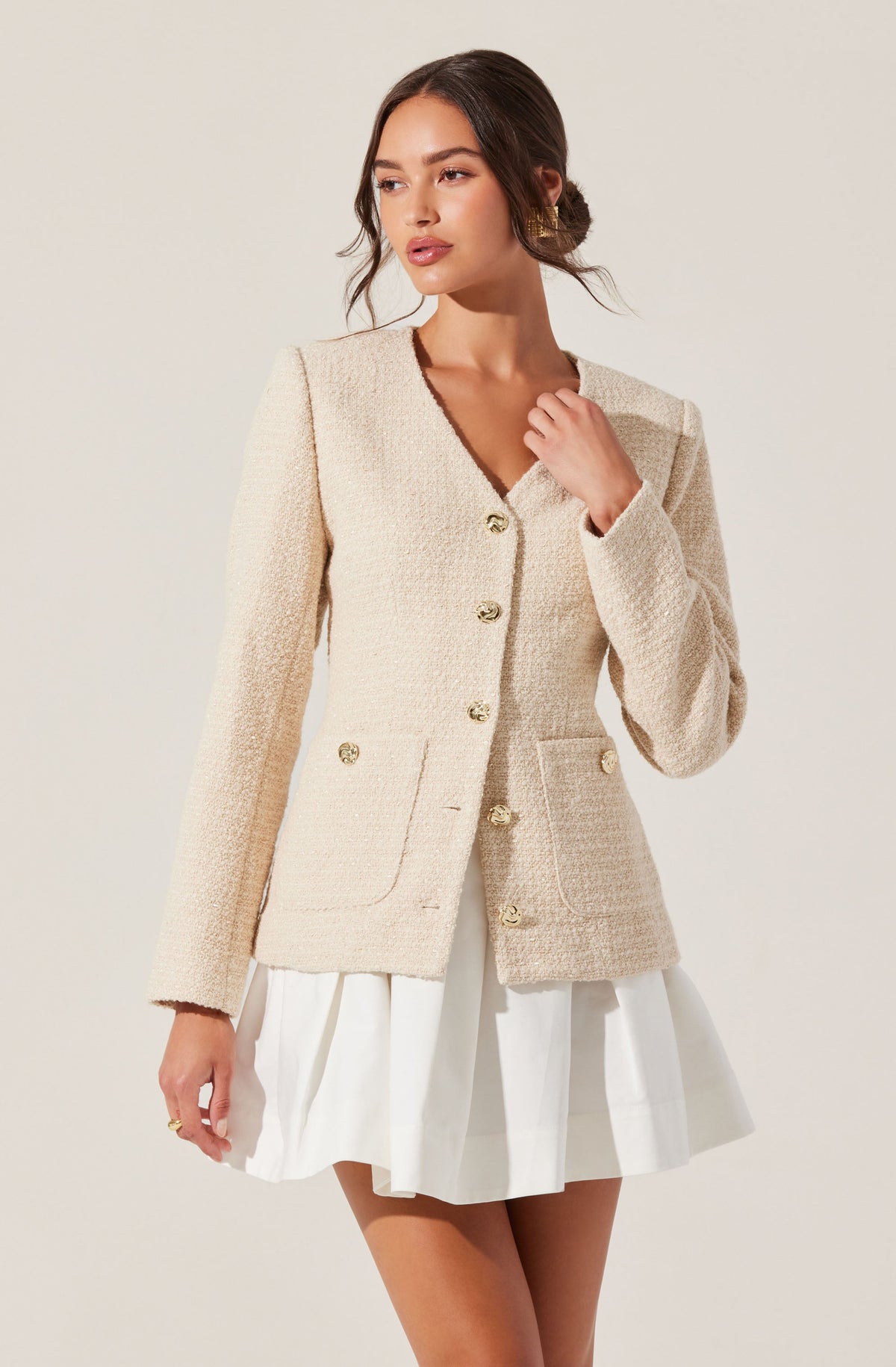 A woman is wearing a cream-colored, textured blazer with gold buttons and two front pockets, paired with a white pleated skirt.