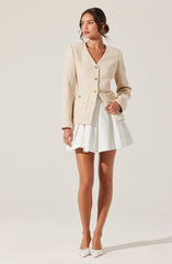 A woman is wearing a cream-colored textured jacket with gold buttons, paired with a white pleated skirt. She has white flat shoes with bows and is standing against a plain light background.