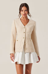 A woman is wearing an elegant cream-colored textured blazer with gold buttons down the front and on the pockets. The blazer has a tailored fit with a V-neckline. She pairs it with a white pleated skirt.