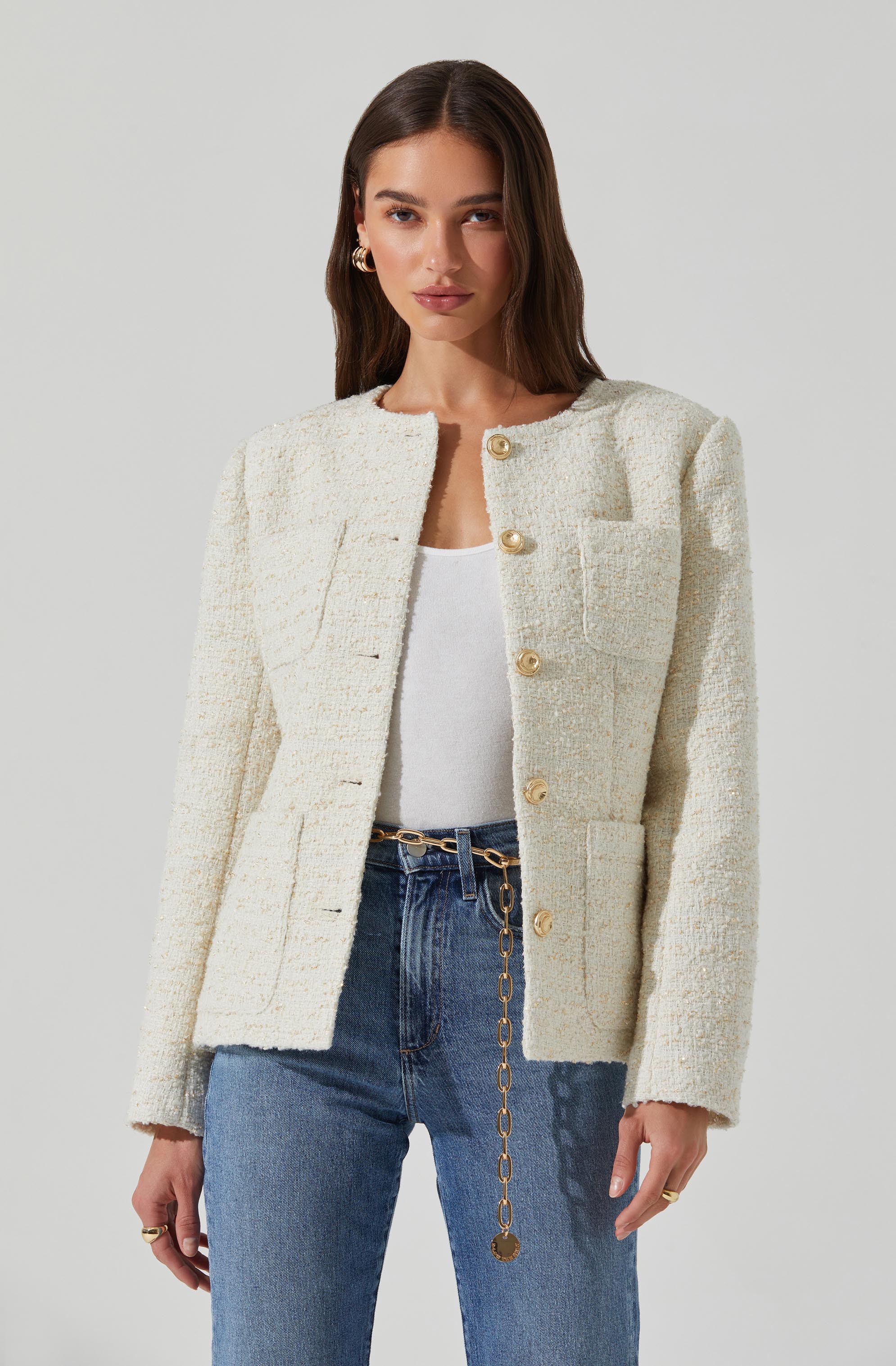 A woman is wearing a cream-colored, textured, cropped blazer with large, gold buttons down the front. The blazer has structured shoulders and patch pockets on the chest. She pairs it with a white top and high-waisted blue jeans, accessorized with a gold chain belt and statement earrings.