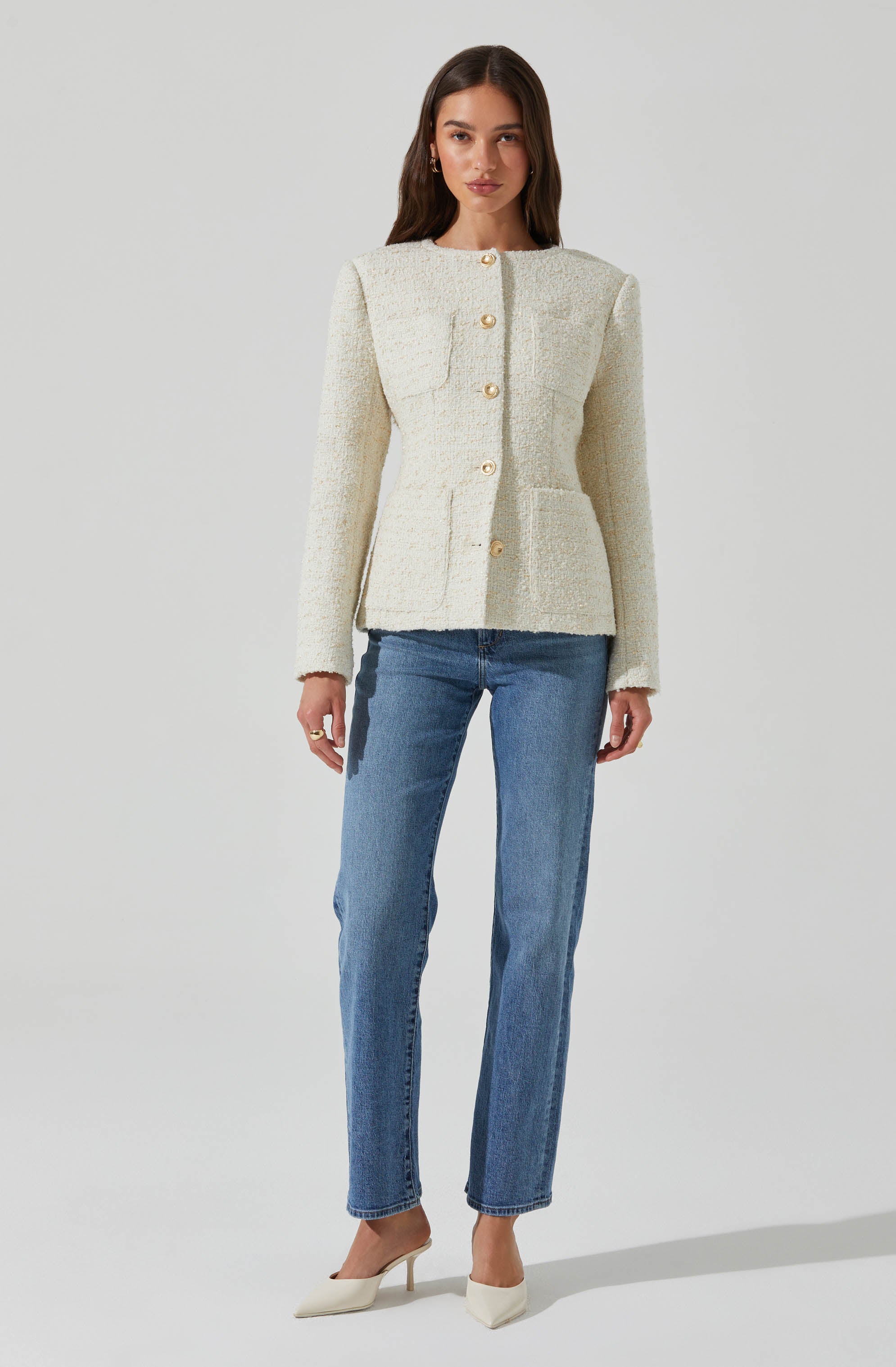 A woman is wearing a cream-colored textured blazer with a round neckline and large gold buttons down the front. The blazer has structured shoulders and two front patch pockets. She pairs it with high-waisted, light-wash straight-leg jeans and white pointed-toe heels.