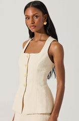 A woman is wearing a sleeveless, cream-colored textured dress with a square neckline and large buttons down the front. The dress has a fitted waist and a slightly flared hem. She accessorizes with gold statement earrings.