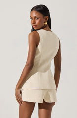 A woman is wearing a sleeveless, cream-colored textured top with a fitted waist and a peplum hem. She is also wearing matching textured shorts. The fabric appears to be a boucle or tweed-like material. She has gold circular earrings and a ring on her left hand.